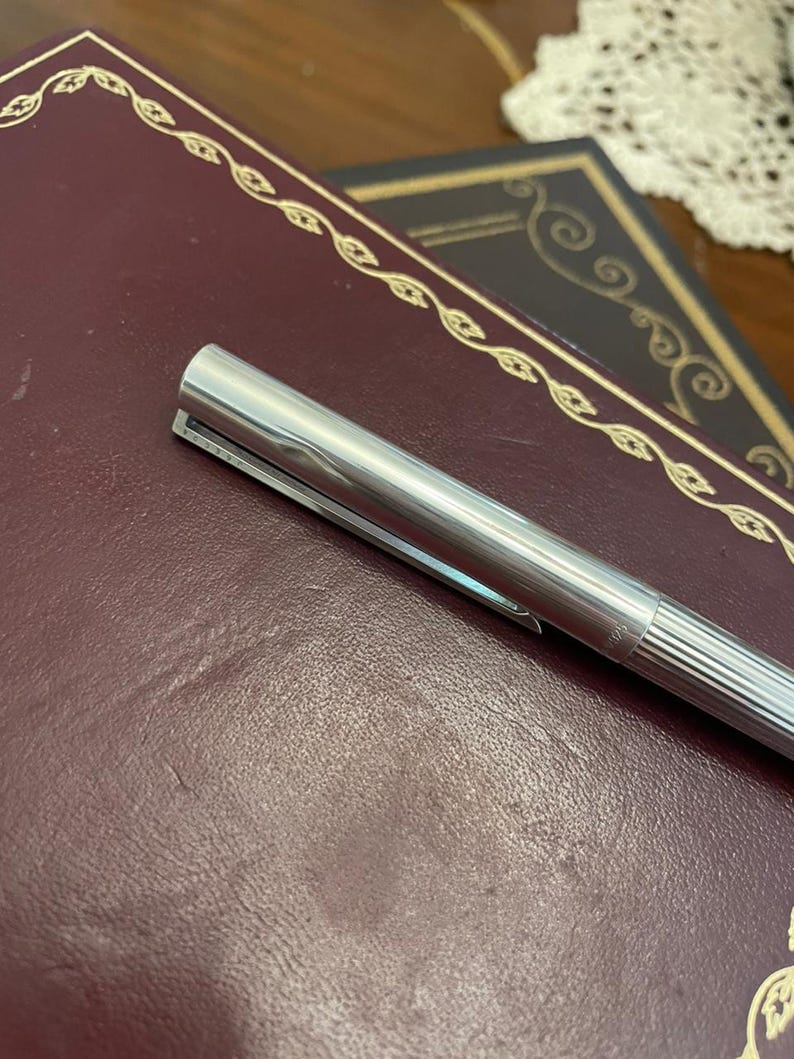 Puede incluir: Un bol&iacute;grafo plateado con clip descansa sobre un libro encuadernado en cuero burdeos. El bol&iacute;grafo tiene un dise&ntilde;o acanalado en la parte inferior del cuerpo y un acabado met&aacute;lico brillante. El libro tiene adornos dorados.