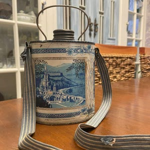 Puede incluir: Contenedor metálico vintage con un diseño de paisaje azul y blanco, con un castillo y un puente. Tiene un asa metálica y una correa para el hombro a rayas azules y blancas. El contenedor es cilíndrico y parece ser una cantimplora o un artículo de picnic.