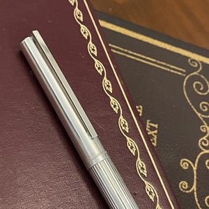 Puede incluir: Un bol&iacute;grafo plateado con un cuerpo acanalado y un clip, apoyado sobre un libro encuadernado en cuero burdeos. El bol&iacute;grafo tiene grabado "S.T. Dupont". El libro tiene un borde decorativo dorado.