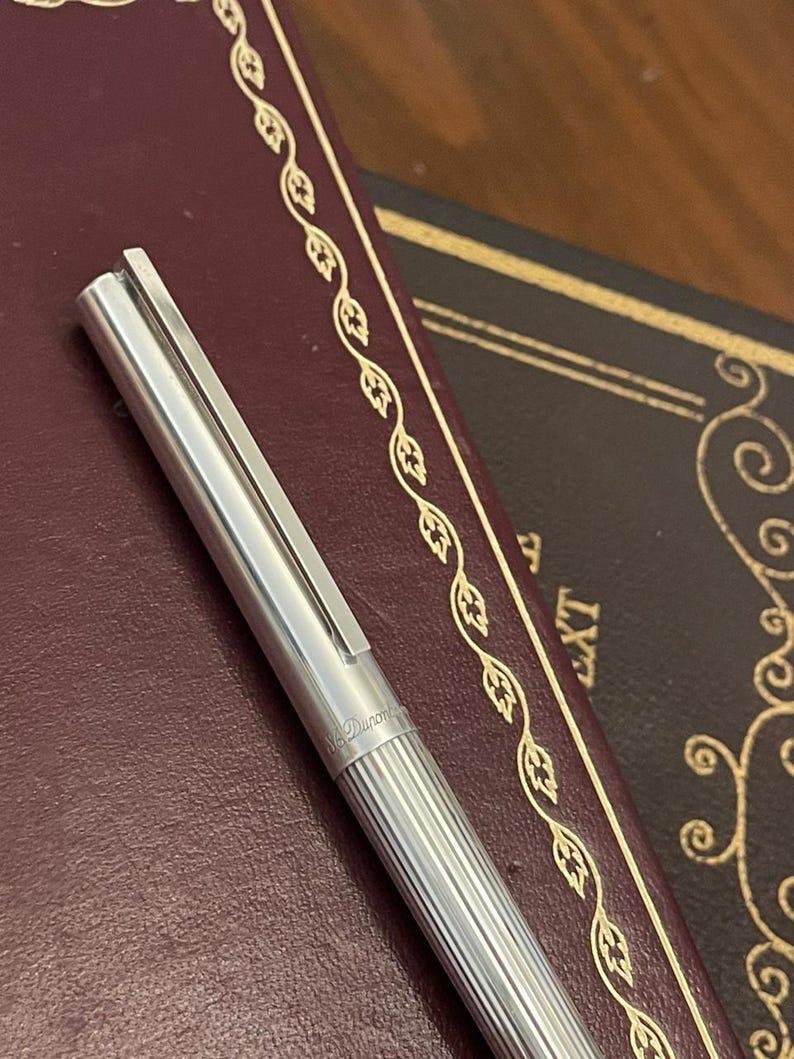 Puede incluir: Un bol&iacute;grafo plateado con un cuerpo acanalado y un clip, apoyado sobre un libro encuadernado en cuero burdeos. El bol&iacute;grafo tiene grabado "S.T. Dupont". El libro tiene un borde decorativo dorado.