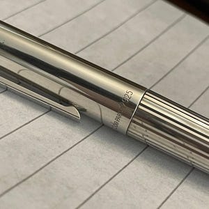 Puede incluir: Primer plano de un bol&iacute;grafo plateado con clip y cuerpo acanalado. El bol&iacute;grafo tiene el texto "de PARIS 925" grabado en el lateral. El bol&iacute;grafo est&aacute; sobre papel rayado.