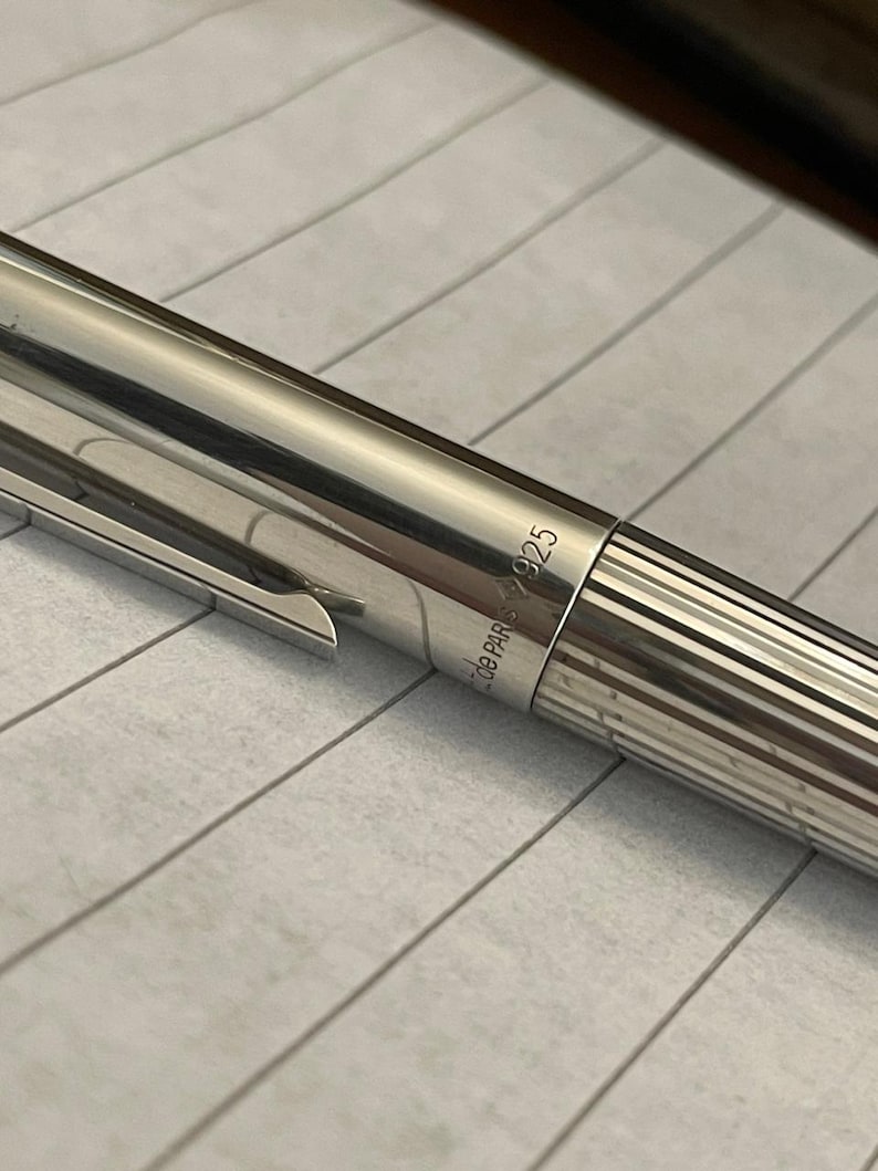 Puede incluir: Primer plano de un bol&iacute;grafo plateado con clip y cuerpo acanalado. El bol&iacute;grafo tiene el texto "de PARIS 925" grabado en el lateral. El bol&iacute;grafo est&aacute; sobre papel rayado.