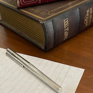 Puede incluir: Un bol&iacute;grafo plateado con dise&ntilde;o acanalado descansa sobre papel rayado, con dos libros antiguos en el fondo. Los libros son de color marr&oacute;n oscuro y burdeos, con detalles dorados y el texto "SAMPLE TEXT" visible.