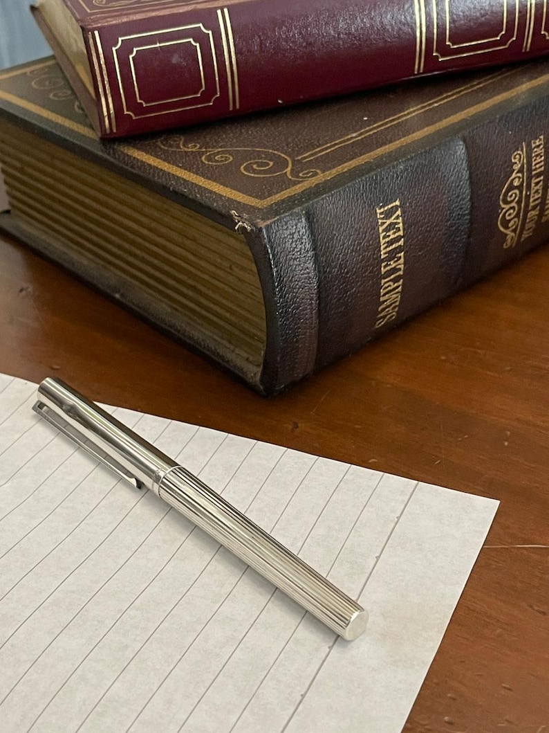 Puede incluir: Un bol&iacute;grafo plateado con dise&ntilde;o acanalado descansa sobre papel rayado, con dos libros antiguos en el fondo. Los libros son de color marr&oacute;n oscuro y burdeos, con detalles dorados y el texto "SAMPLE TEXT" visible.