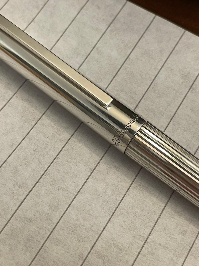 Puede incluir: Primer plano de un bol&iacute;grafo plateado con una superficie brillante y reflectante. El bol&iacute;grafo tiene un clip rectangular y un dise&ntilde;o a rayas en el cuerpo inferior. La marca "S.T. Dupont" est&aacute; grabada en el bol&iacute;grafo.