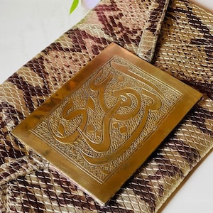 May include: A snakeskin-patterned clutch in shades of brown and tan, with a rectangular, gold-toned metal plate featuring ornate calligraphy. The clutch is folded over, and the metal plate is centered on the flap.