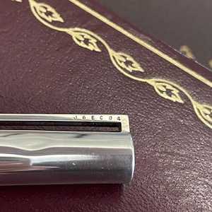 Puede incluir: Primer plano de un objeto met&aacute;lico plateado brillante con el texto "J6EC04" grabado en la parte superior. El objeto se encuentra sobre un fondo oscuro y texturizado con adornos decorativos dorados.