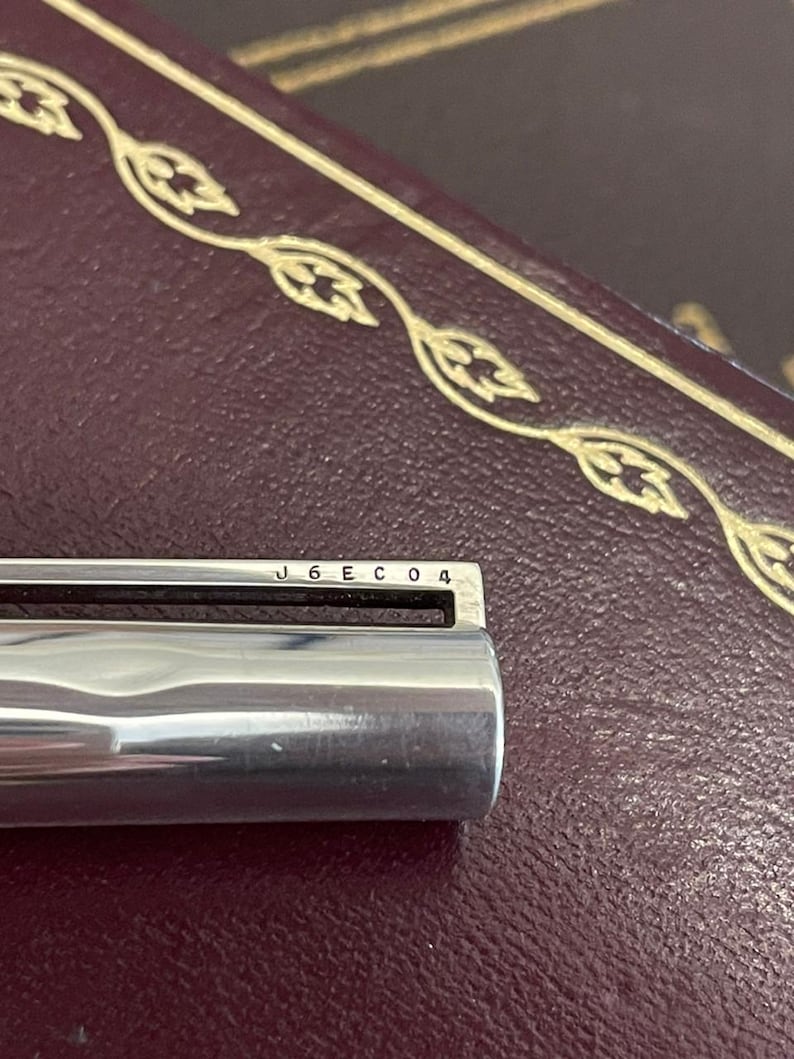 Puede incluir: Primer plano de un objeto met&aacute;lico plateado brillante con el texto "J6EC04" grabado en la parte superior. El objeto se encuentra sobre un fondo oscuro y texturizado con adornos decorativos dorados.