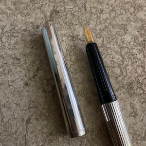 Puede incluir: Una pluma estilogr&aacute;fica plateada y negra con plum&iacute;n dorado, la tapa est&aacute; quitada y colocada junto a la pluma. La pluma tiene un dise&ntilde;o acanalado en el cuerpo y el capuch&oacute;n. El plum&iacute;n es de color dorado.