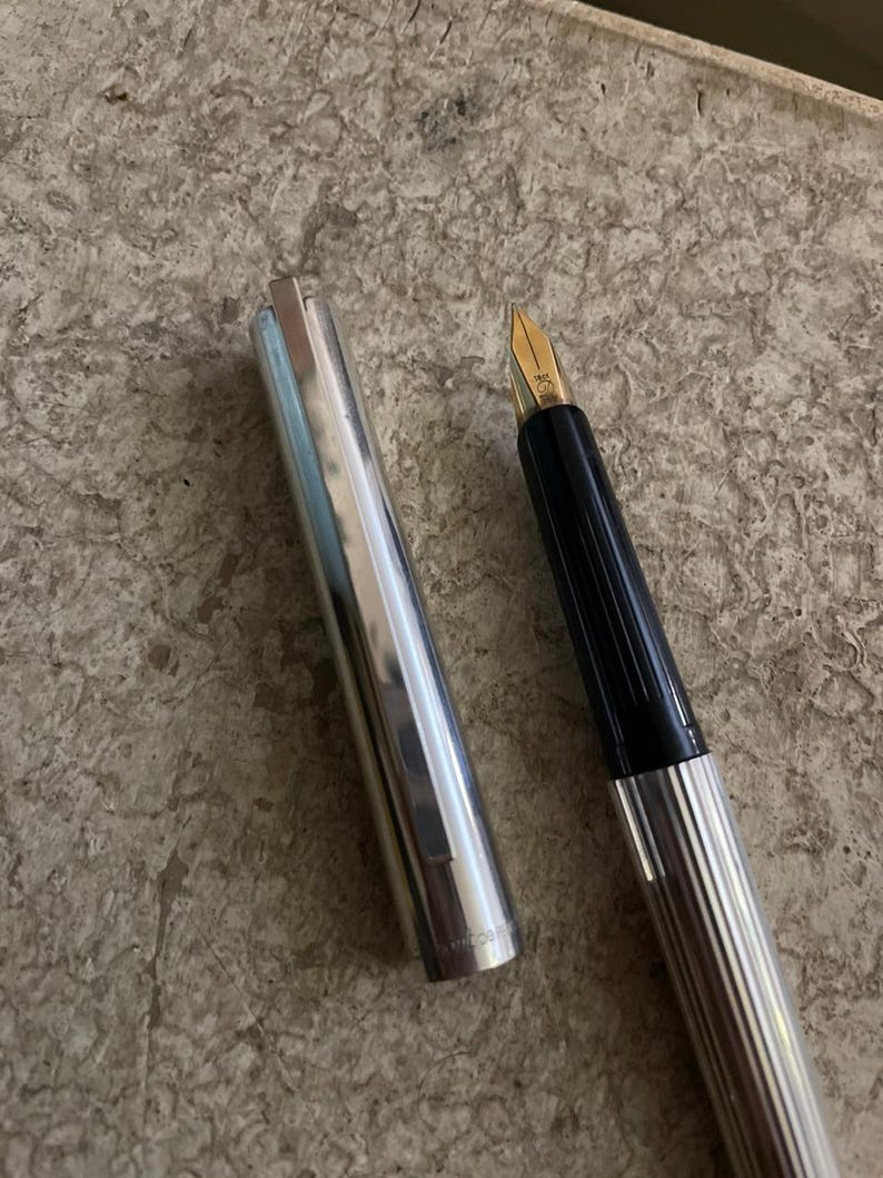 Puede incluir: Una pluma estilogr&aacute;fica plateada y negra con plum&iacute;n dorado, la tapa est&aacute; quitada y colocada junto a la pluma. La pluma tiene un dise&ntilde;o acanalado en el cuerpo y el capuch&oacute;n. El plum&iacute;n es de color dorado.