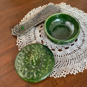 Puede incluir: Un cuenco de cerámica verde con tapa a juego y un portaincienso decorativo plateado. El cuenco y la tapa son de color verde oscuro brillante. El portaincienso es plateado con intrincados diseños y una gema verde.