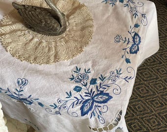 Könnte beinhalten: Weißes Leinentischtuch mit blauer Blumenstickerei. Eine silberne Schwanfigur liegt auf einer cremefarbenen Spitzendecke. Das Tischtuch hat Ausschnittdetails am Rand, was eine zarte und elegante Ästhetik erzeugt.