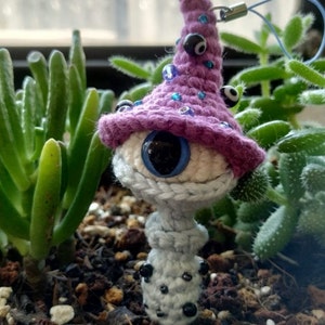 Puede incluir: Un hongo de crochet con un sombrero morado y un gran ojo azul. El sombrero está decorado con cuentas azules y negras. El hongo está unido a una cuerda negra.