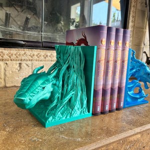 Puede incluir: Un par de sujetalibros de color verde azulado con tem&aacute;tica de drag&oacute;n que sostienen cuatro libros. El sujetalibros izquierdo presenta una cabeza de drag&oacute;n detallada que emerge de una estructura texturizada. El derecho muestra un drag&oacute;n.
