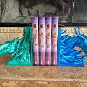 Puede incluir: Un par de sujetalibros con forma de drag&oacute;n que sostienen cuatro copias del libro Wings of Fire: The Hidden Prince. El sujetalibros izquierdo es verde met&aacute;lico y el derecho es azul met&aacute;lico. Ambos presentan cabezas de drag&oacute;n detalladas.