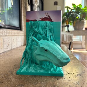 Puede incluir: Un sujetalibros de color verde azulado impreso en 3D que presenta una cabeza de drag&oacute;n esculpida que emerge de una estructura texturizada similar a ra&iacute;ces. El drag&oacute;n tiene cuernos y escamas visibles, y la base es rectangular.