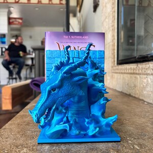 Puede incluir: Un sujetalibros de drag&oacute;n azul brillante impreso en 3D con un patr&oacute;n de escamas texturizado, cuernos curvos y una base en forma de llama. El drag&oacute;n est&aacute; colocado frente a un libro titulado Wings of Fire de Tui T. Sutherland con un muro de ladrillos.