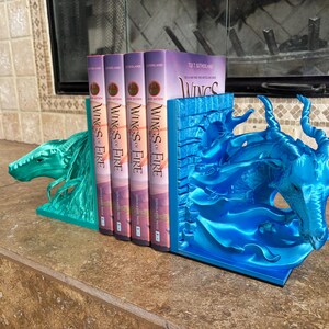 Puede incluir: Un par de sujetalibros de drag&oacute;n azul met&aacute;lico que sostienen cuatro libros de Wings of Fire. El sujetalibros izquierdo presenta una cabeza de drag&oacute;n mirando a la izquierda, mientras que el derecho muestra una cabeza de drag&oacute;n mirando a la derecha.
