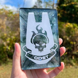 Peut inclure: Un objet rectangulaire en résine bleu sarcelle et noire avec un graphisme blanc d'un crâne avec des flammes, une lettre A stylisée et le texte "ODST". Le fond a un effet marbré.