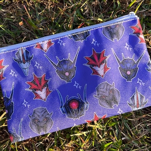 May include: A rectangular zipped pouch featuring a blue background with a repeating pattern of robot face illustrations in various colours. The pouch is on green grass.
