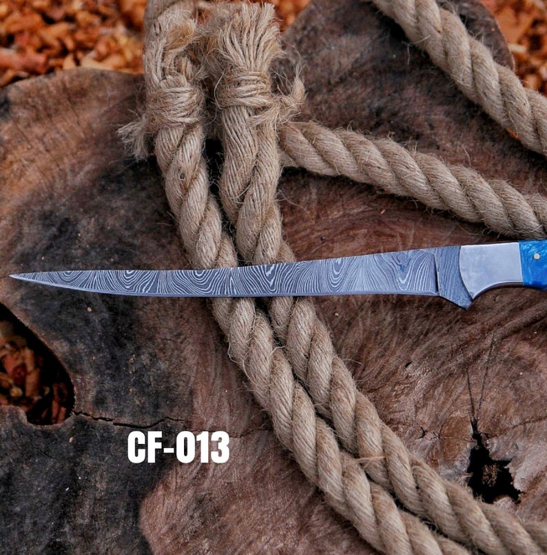 May include: A long, patterned steel knife with a blue handle, resting on a wooden surface with a thick rope. The blade has a unique wave-like pattern. The text "CF-013" is displayed in white.