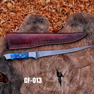 May include: A long, stainless steel knife with a blue handle and a brown leather sheath. The knife blade has a unique pattern. The handle has a blue and silver design. The sheath is brown with decorative stitching. The text "CF-013" is visible.