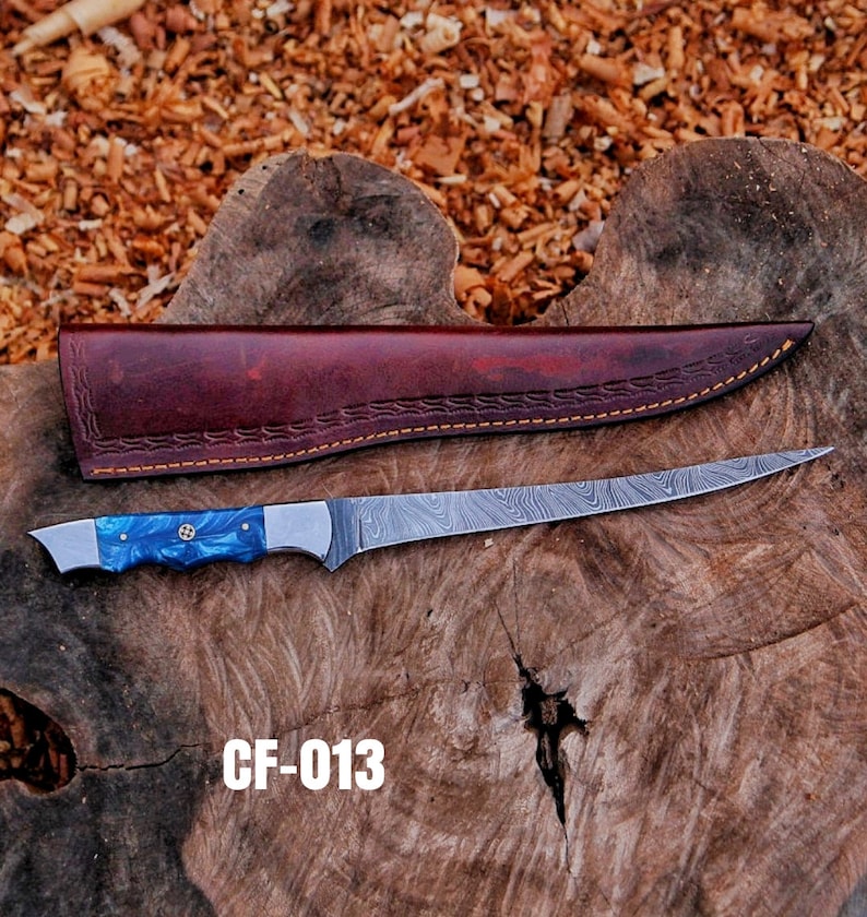 May include: A long, stainless steel knife with a blue handle and a brown leather sheath. The knife blade has a unique pattern. The handle has a blue and silver design. The sheath is brown with decorative stitching. The text "CF-013" is visible.