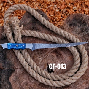 May include: A long, stainless steel knife with a blue handle and a patterned blade. The handle has a decorative silver emblem. The knife is displayed on a wooden surface with a coiled rope. The text "CF-013" is visible.