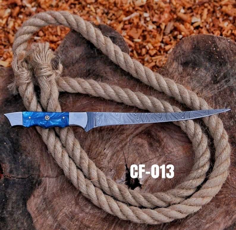 May include: A long, stainless steel knife with a blue handle and a patterned blade. The handle has a decorative silver emblem. The knife is displayed on a wooden surface with a coiled rope. The text "CF-013" is visible.