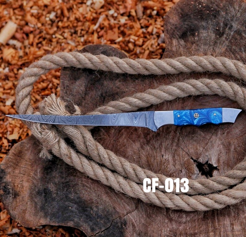 May include: A Damascus steel fillet knife with a blue and white handle, resting on a wooden surface with a coiled rope. The knife has a long, thin blade and the text "CF-013" is visible.