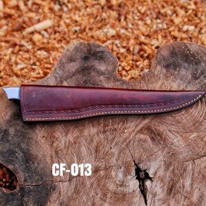 May include: A burgundy leather knife sheath with a decorative stitched pattern and a stainless steel blade. The sheath is marked with the text "CF-013" in white. The knife is resting on a wooden surface.