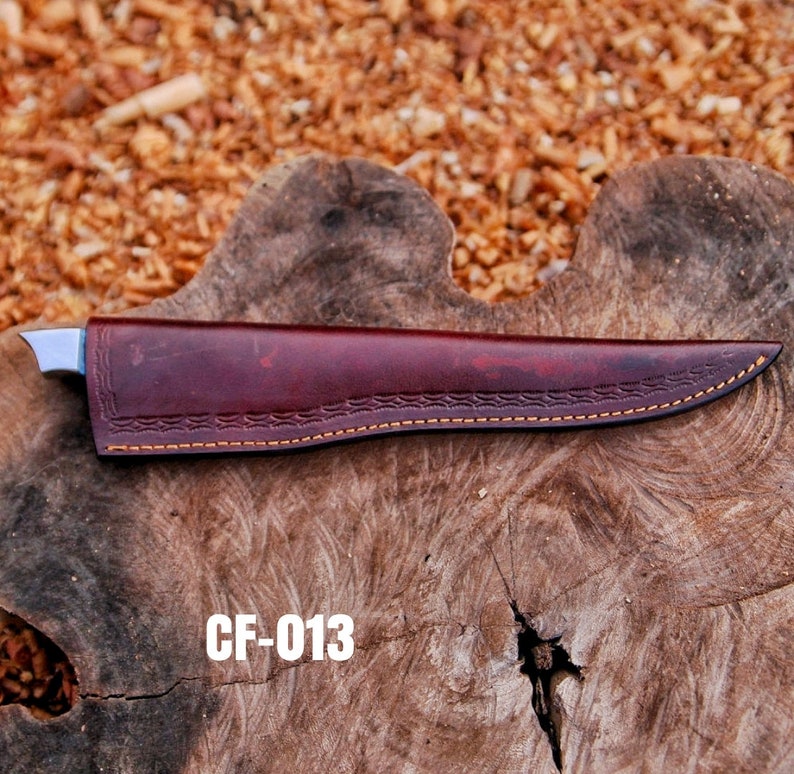 May include: A burgundy leather knife sheath with a decorative stitched pattern and a stainless steel blade. The sheath is marked with the text "CF-013" in white. The knife is resting on a wooden surface.