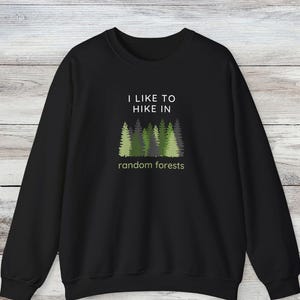 Könnte beinhalten: Ein schwarzes Langarm-Sweatshirt mit einer Grafik aus verschiedenen grünen und grauen Tannenbäumen. Über den Bäumen steht der Text I LIKE TO HIKE IN in Weiß, und unter den Bäumen steht der Text random forests in einer hellgrünen Farbe.