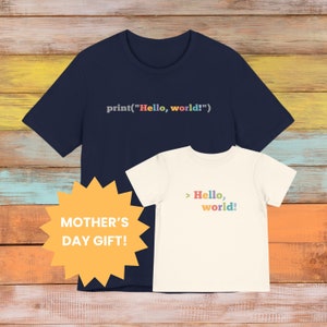May include: Two t-shirts, one navy blue and one white, with the text "print("Hello, world!")" and "> Hello, world!" printed on them. The shirts are on a wooden background with a yellow starburst sticker that says "MOTHER'S DAY GIFT!"