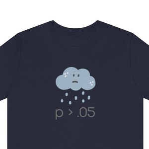 May include: Navy blue t-shirt featuring a sad cloud graphic with raindrops and the text "p > .05". The cloud is light blue with a small face. The text is in a gray color. The shirt is made of a soft material.
