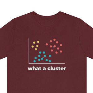 May include: Maroon t-shirt featuring a data visualization graphic with yellow, red, and blue dots, and the text "what a cluster" in white. The graphic resembles a scatter plot, with dots grouped together. The shirt is made of a soft material.