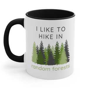 May include: White ceramic mug with a black rim and handle. The mug has a green and black forest design and the text "I like to hike in random forests".