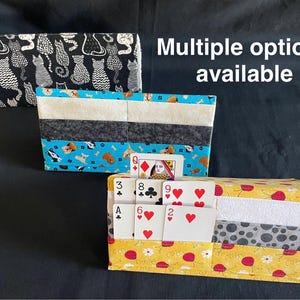 Puede incluir: Tres tarjeteros de tela con diferentes estampados. Uno es negro con siluetas de gatos blancos, otro tiene un estampado azul de perros y huesos, y el tercero es amarillo con detalles de flores rojas. Hay naipes en el tarjetero amarillo. Texto: Múltiples opciones disponibles.