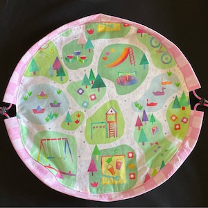 Puede incluir: Una alfombra de juego rosa y blanca con una ilustración colorida de un parque infantil. La alfombra de juego tiene un cierre de cordón y es perfecta para jugar en interiores o exteriores.