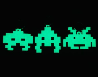 May include: Three glow-in-the-dark keychains shaped like the classic video game alien, Space Invaders. The keychains are green and have a pixelated design.