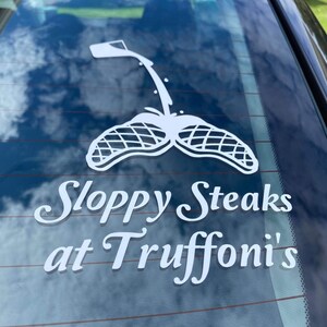 Può includere: Decalcomania bianca su un finestrino dell'auto con un'immagine stilizzata di un contenitore che versa e due forme di bistecca. Il testo recita "Sloppy Steaks at Truffoni's" in un carattere decorativo. Lo sfondo mostra un cielo nuvoloso.