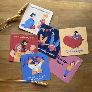 May include: A collection of illustrated square cards with various designs and text in a foreign language. The cards feature illustrations of people, flowers, and hearts, with a rustic, textured appearance. The cards are arranged on a wooden surface.