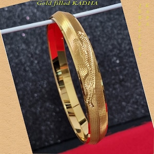 May include: Gold filled KADHAA bangle bracelet with a decorative design. The bangle is a shiny gold color.