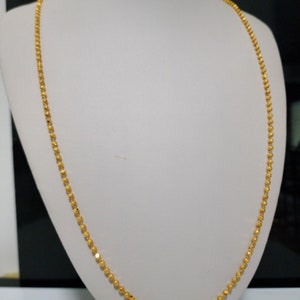 Puede incluir: Un delicado collar dorado con un diseño de cuentas. El collar se exhibe en un maniquí blanco, mostrando su longitud y detalles intrincados. Las cuentas parecen pequeñas y espaciadas uniformemente, creando una cadena continua y elegante.
