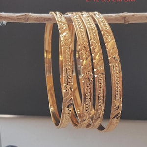 Op de afbeelding: Een set van vijf gouden armbanden met ingewikkelde bloemen- en geometrische patronen. De armbanden worden getoond op een dunne houten stok tegen een donkere achtergrond. Elke armband heeft een diameter van 6,9 cm.