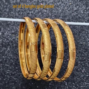 May include: A set of four gold-plated bangles. Each bangle has a textured, repeating pattern. The bangles are displayed on a white rod against a dark, speckled background. The text "set of 4 bangles gold plated" is visible.