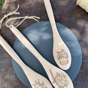 Peut inclure: Trois cuillères en bois avec des motifs gravés sur le thème de l'automne. L'une représente une citrouille et des fleurs, une autre une brouette avec des citrouilles, et la dernière un panier de citrouilles. Chaque cuillère a un trou pour suspendre.