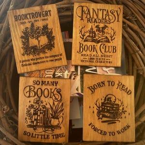 Può includere: Quattro cartelli in legno a tema libro. I cartelli sono decorati con illustrazioni e testo. Il primo cartello recita "Booktrovert [book-trovert/noun] Una persona che preferisce la compagnia di personaggi immaginari alle persone reali". Il secondo cartello recita "Fantasy Readers Book Club Leggi tutta la notte - dipendente dai personaggi immaginari". Il terzo cartello recita "Tanti libri, così poco tempo". Il quarto cartello recita "Nato per leggere, costretto a lavorare".