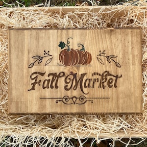 Peut inclure: Panneau rectangulaire en bois avec l'inscription "Fall Market" dans une police décorative. Le panneau présente une illustration de citrouille marron avec des feuilles vertes et des ornements décoratifs. Le panneau est présenté dans une caisse en bois avec de la paille.