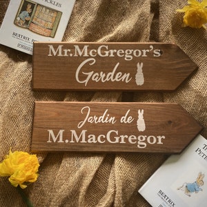 Peut inclure: Deux panneaux en bois en forme de flèche. Le panneau du haut indique "Mr. McGregor's Garden" en lettres blanches avec une silhouette de lapin blanche. Le panneau du bas indique "Jardin de M.MacGregor" en lettres blanches avec une silhouette de lapin blanche.